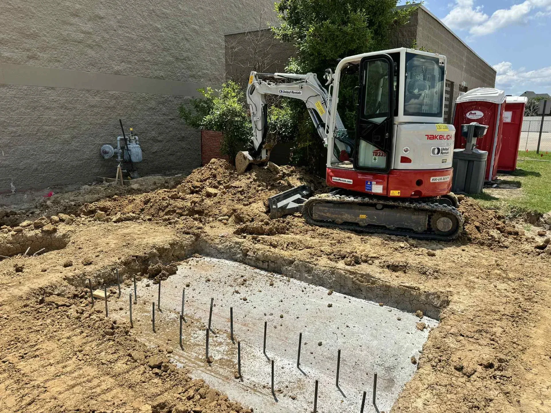 Mini excavator digging in dirt near a concrete foundation. Two portable toilets are in the background.