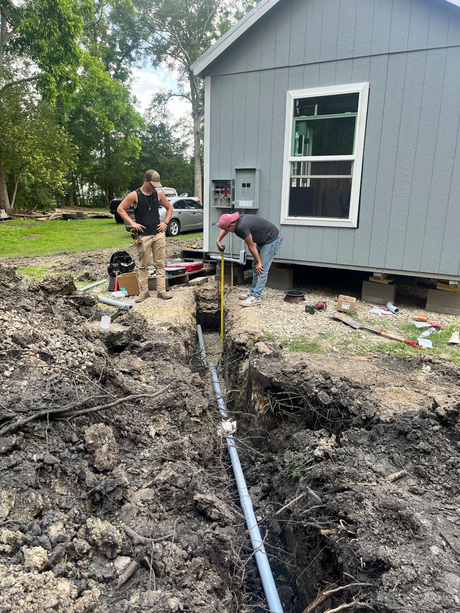 Two workers install plumbing in a trench next to a gray house, one measuring with a rod.