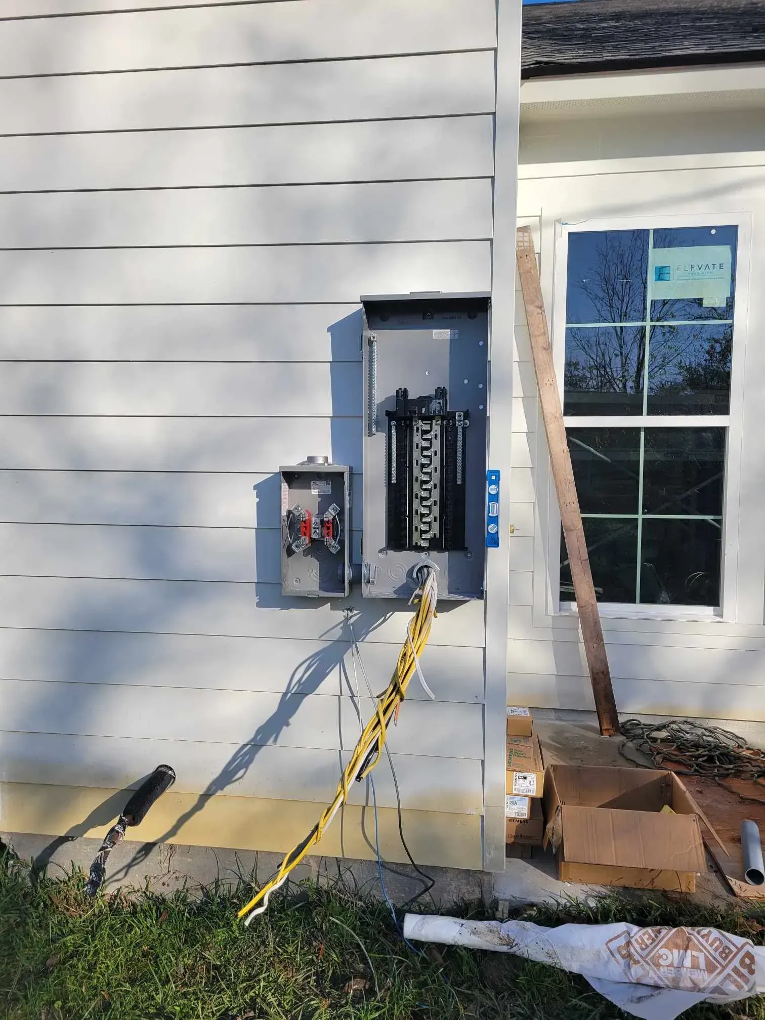Exterior electrical panel mounted on a white house. Wires hang down near grass, boxes and lumber on ground.