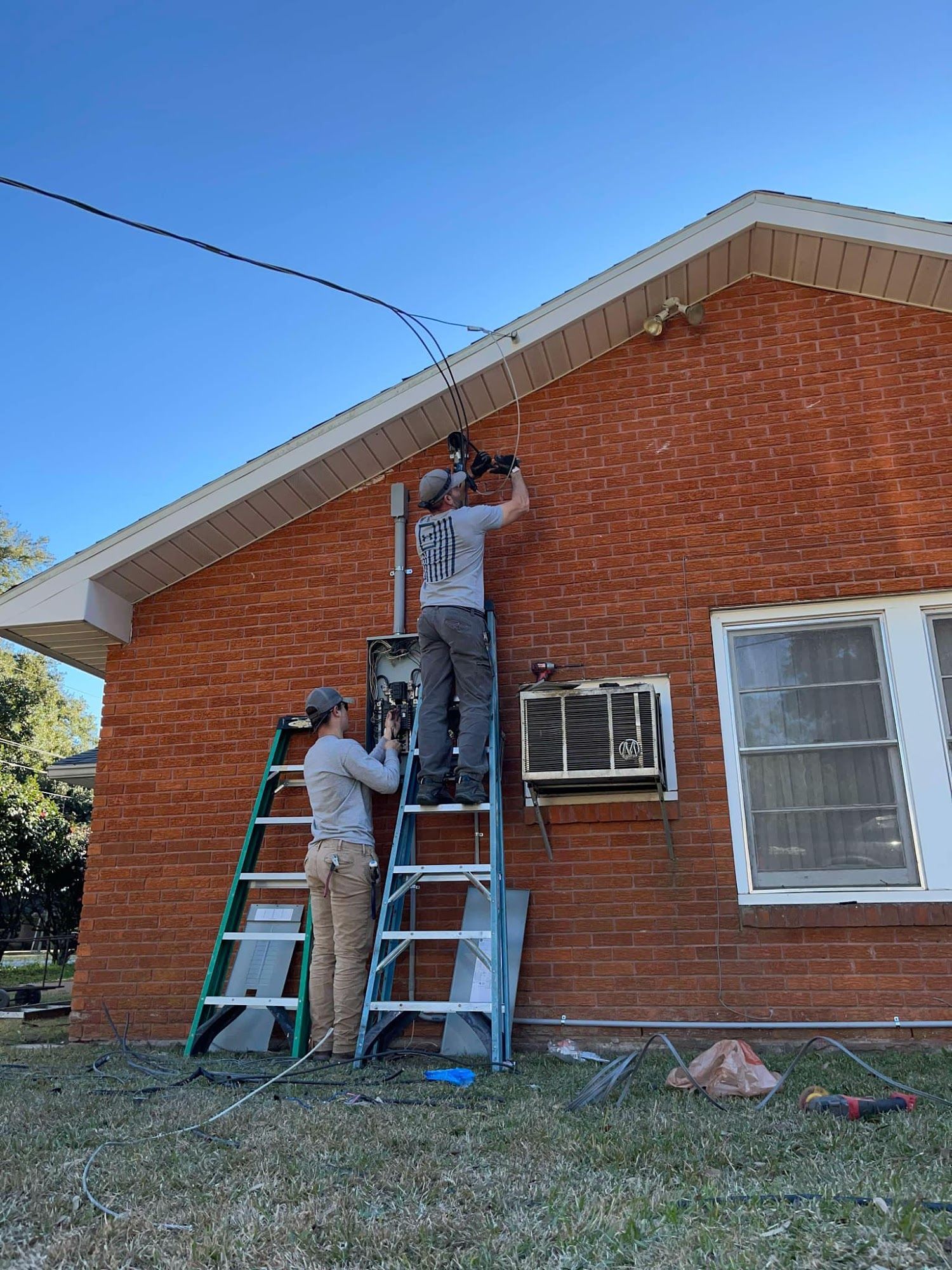 Two workers on ladders installing electrical equipment on a brick building under a clear sky.