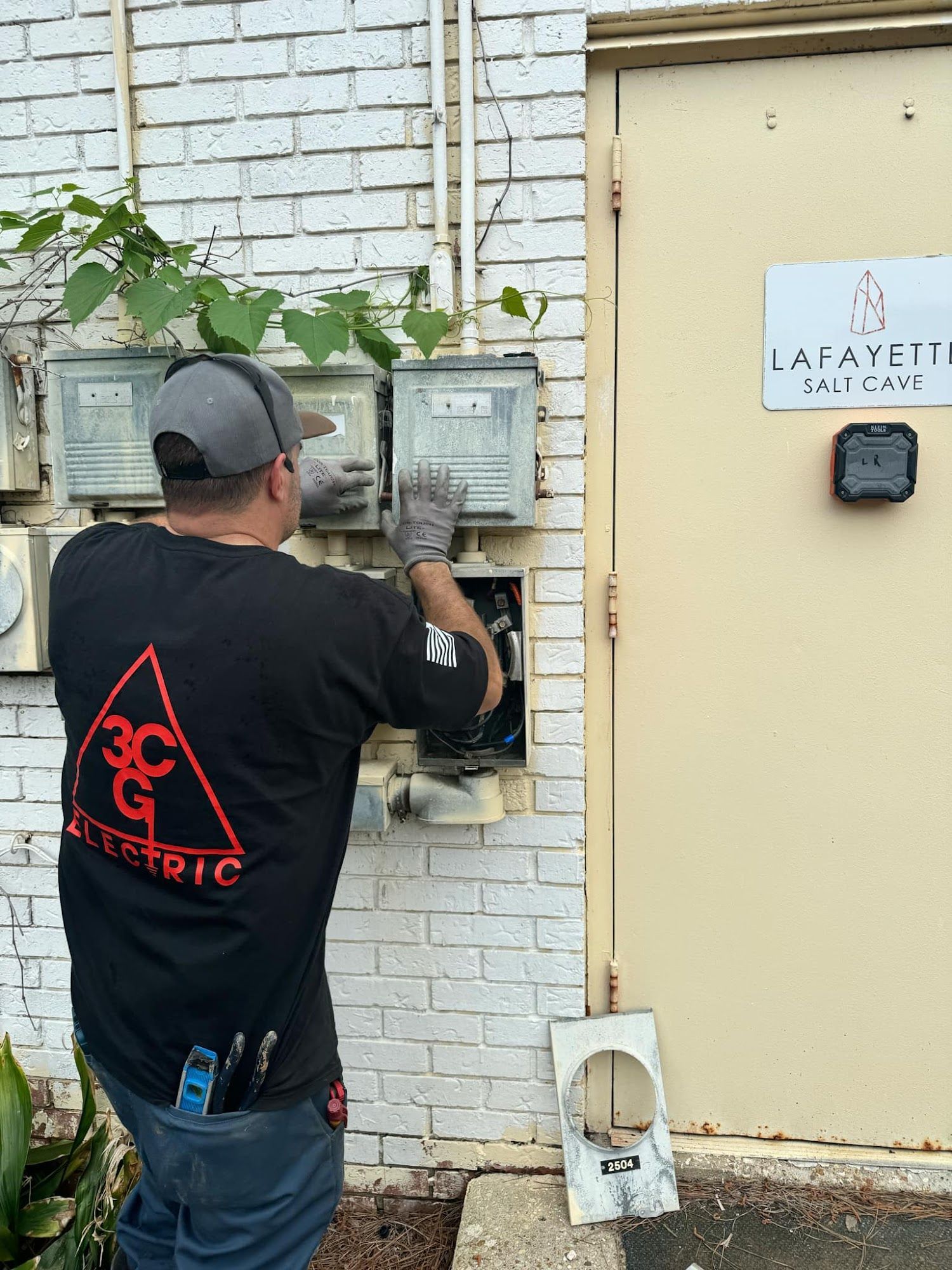 Electrician in grey cap and gloves working on electrical meters on a brick wall. Next to a tan utility door.