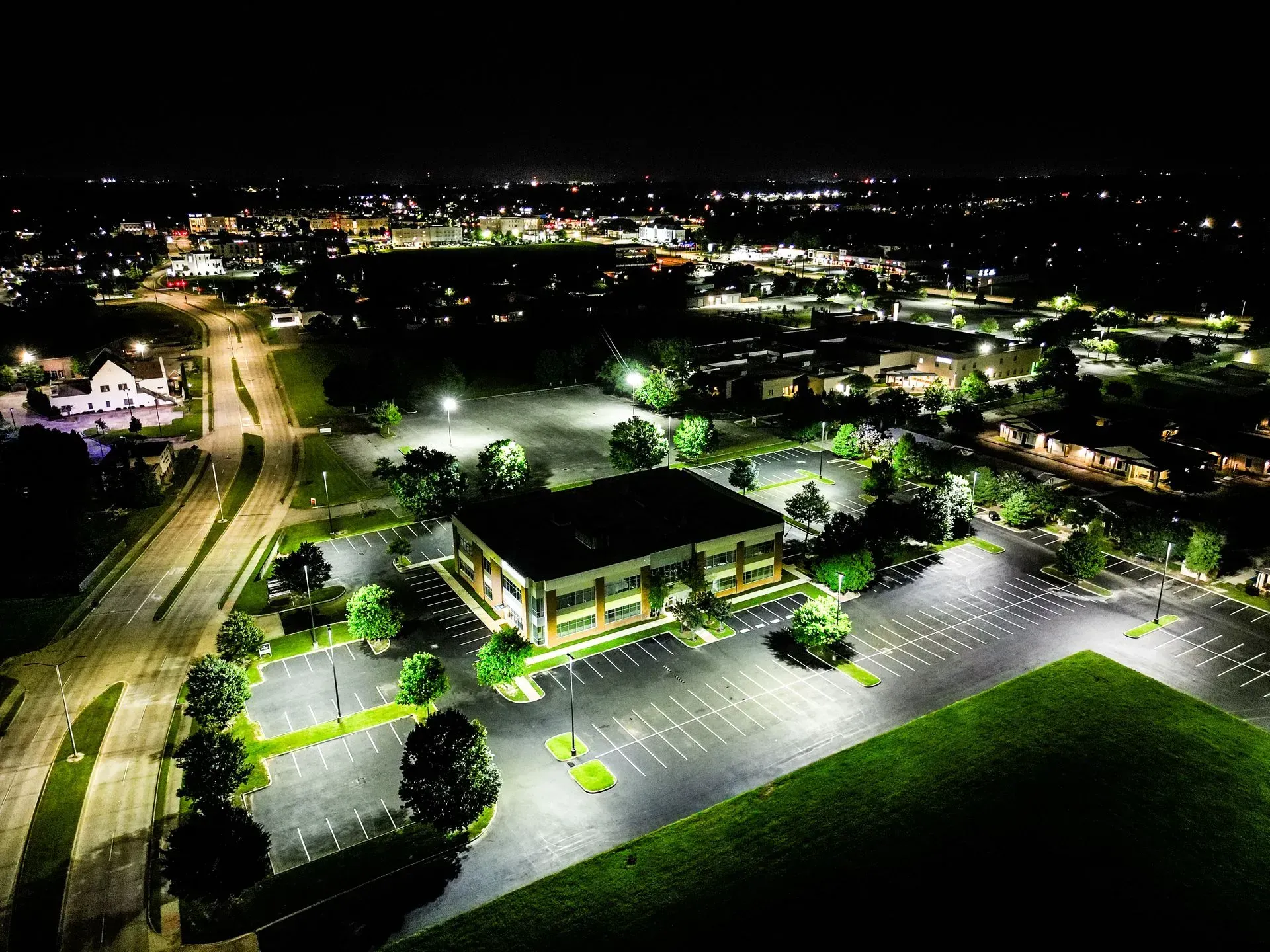 Night aerial view of a building with lit parking lot and distant city lights. Green lawn visible.