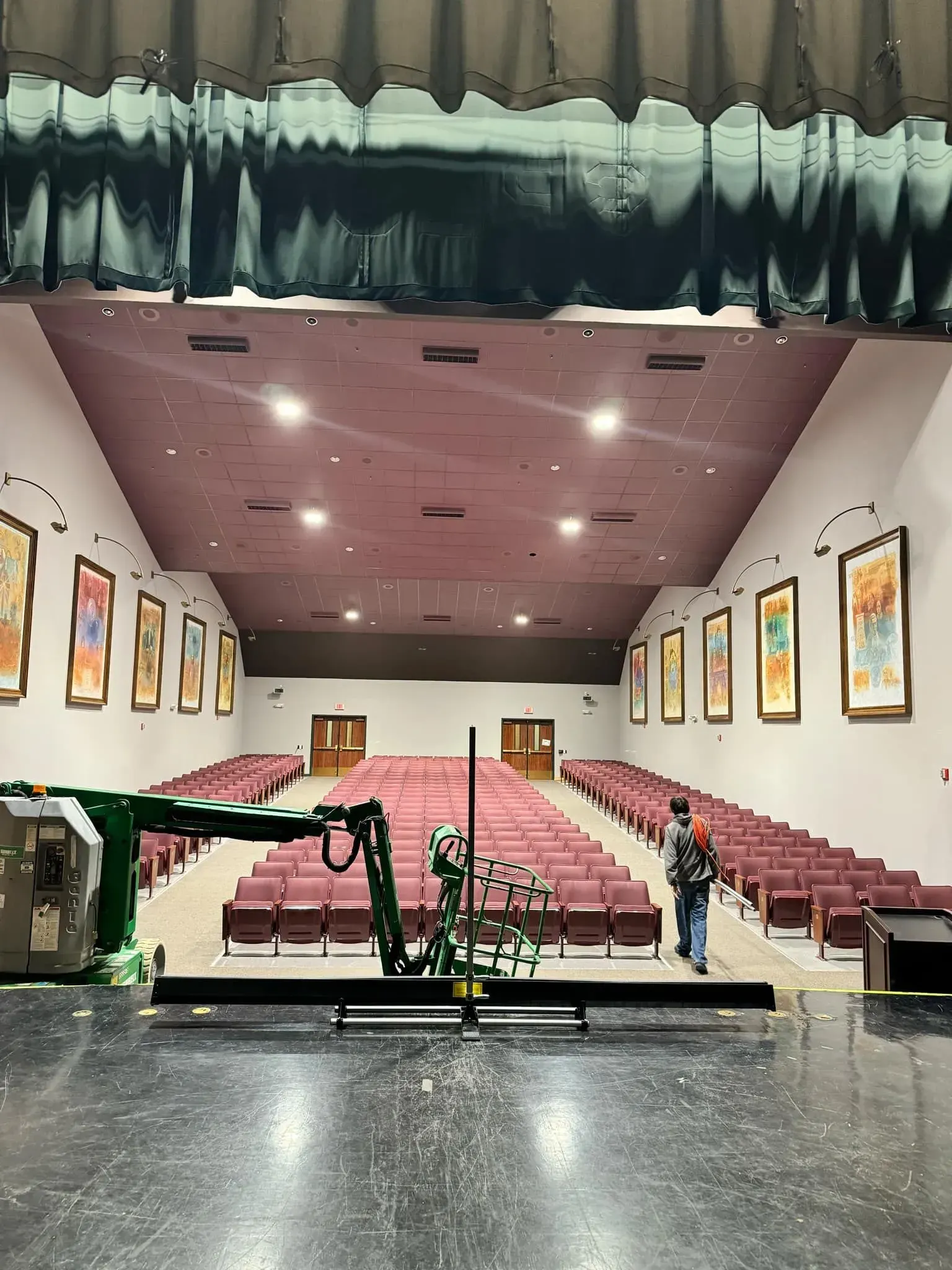 Stage view of a burgundy-seated auditorium. A person walks the aisle. Stage decorations include flowers and a green boom.