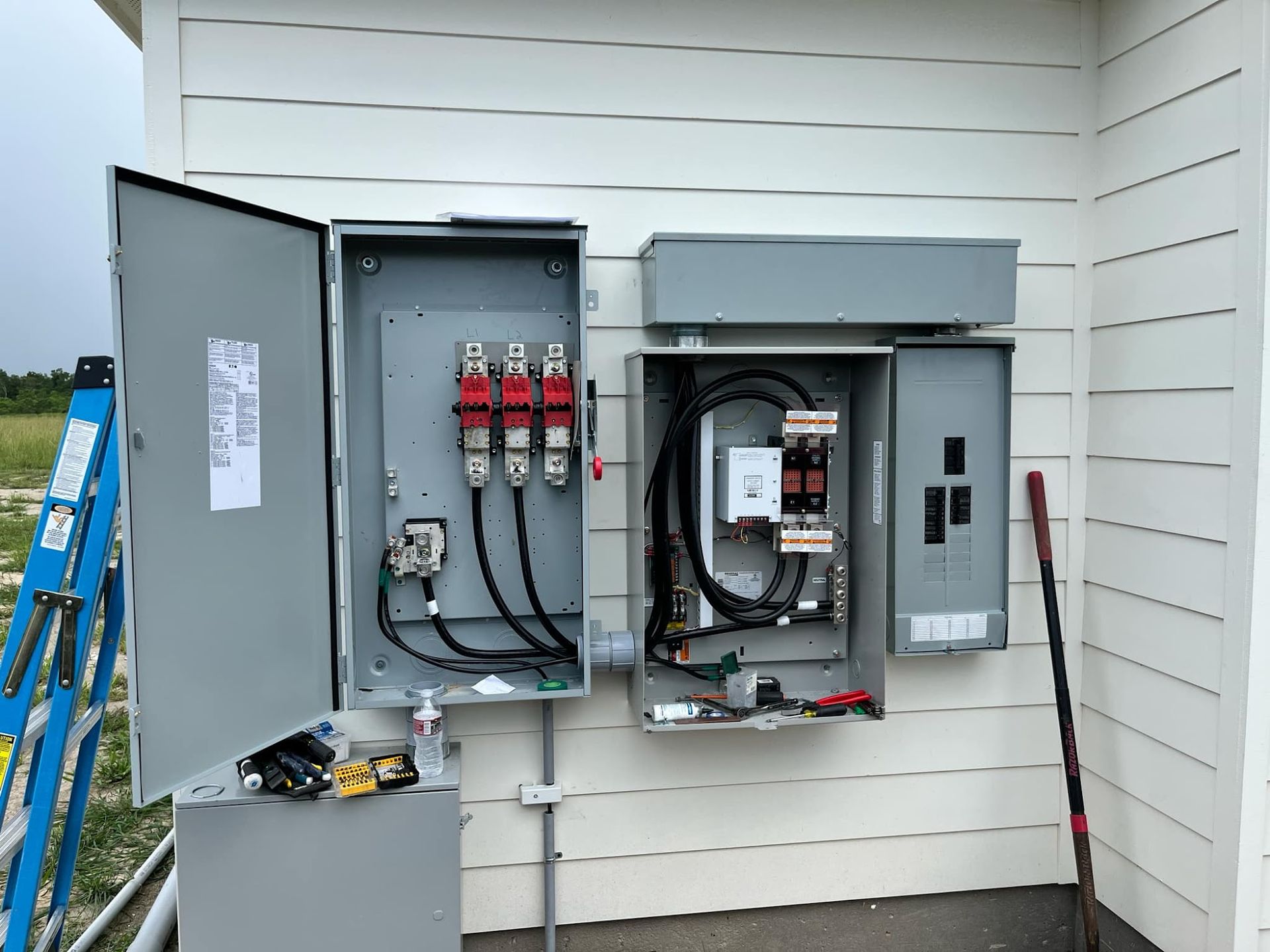 Electrical boxes mounted on a white house exterior with open panels, showing wires and components.