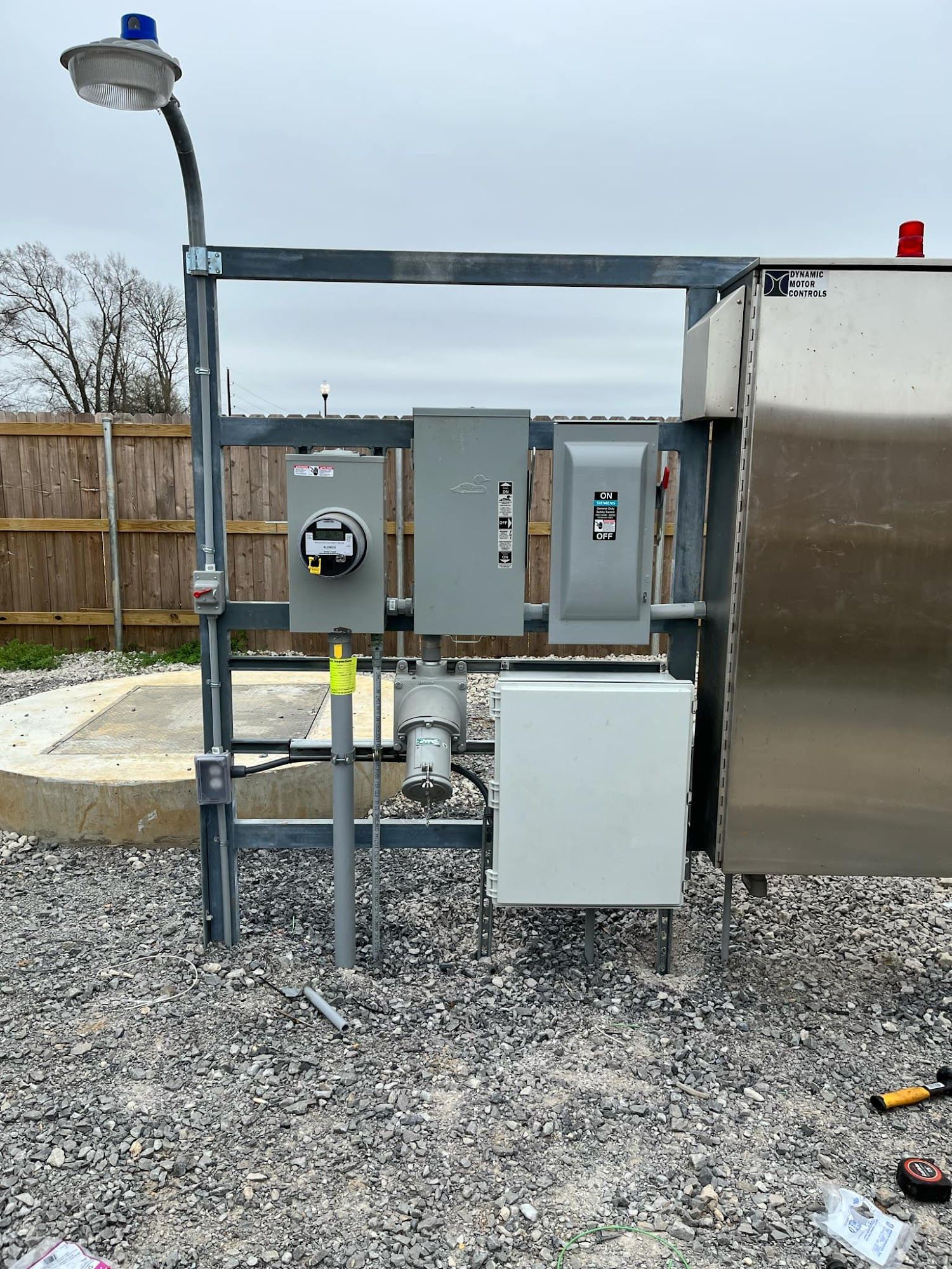 Electrical panel with meter, gray metal frame, light, and stainless steel enclosure on gravel.
