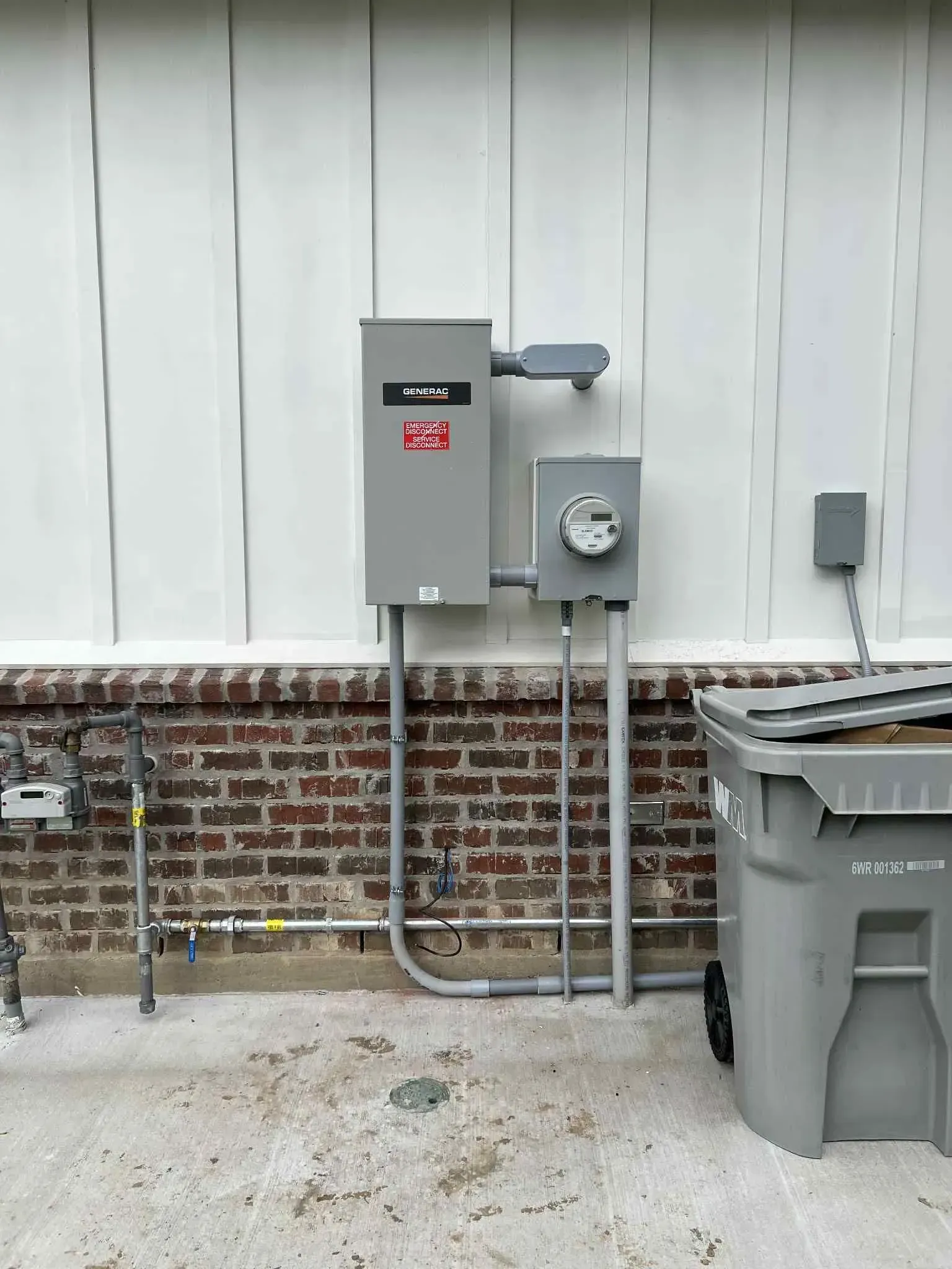 Electrical equipment on a brick and white wall. Includes a meter, panel, and conduit. A trash can is to the right.