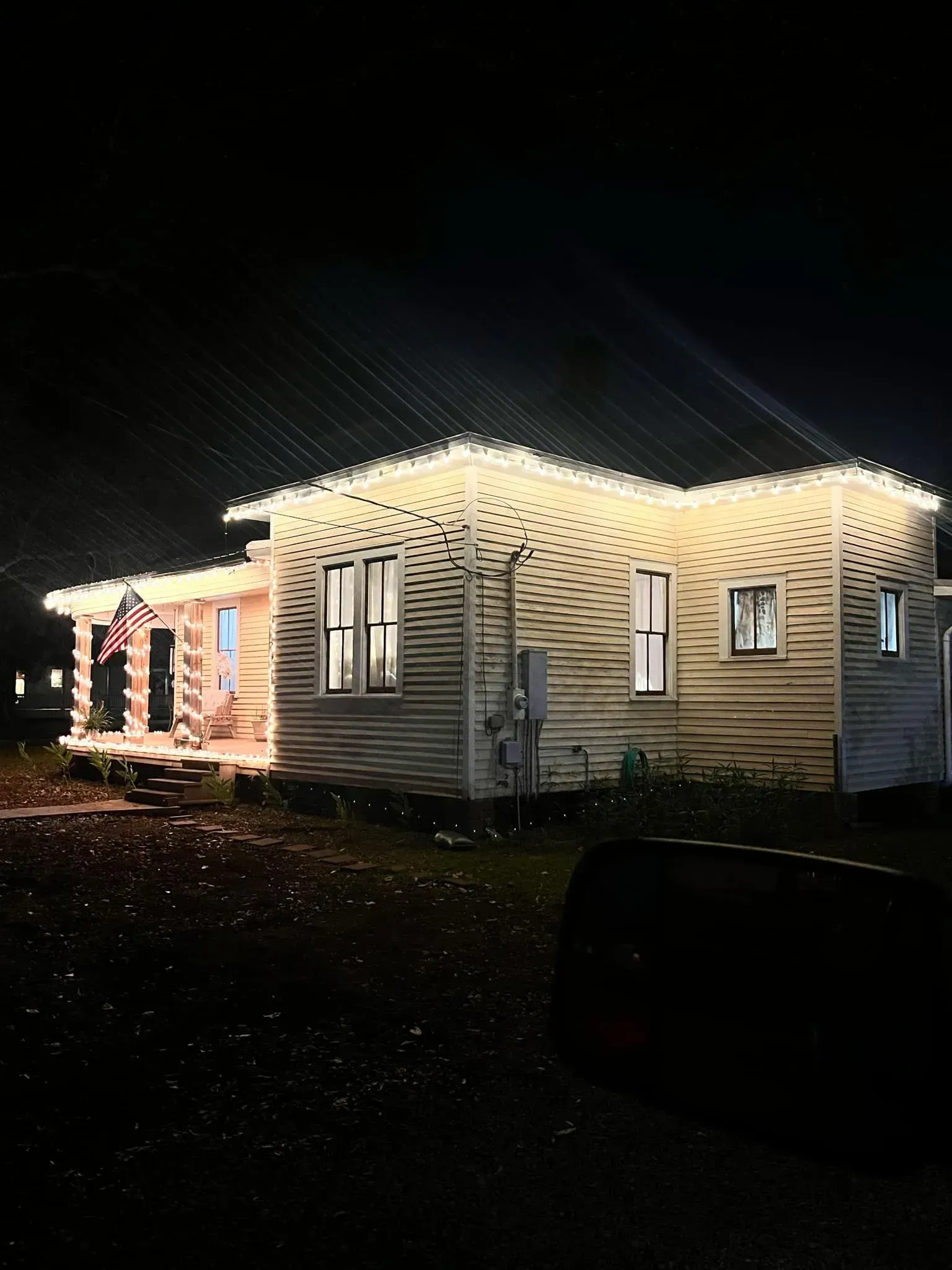 Cottage illuminated with white lights at night; an American flag hangs on the porch.
