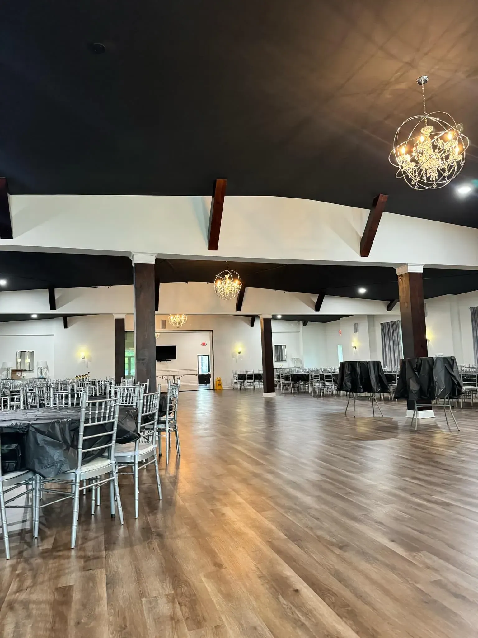 Empty event hall with black ceiling, wood floor, tables covered in black cloth, chandeliers.