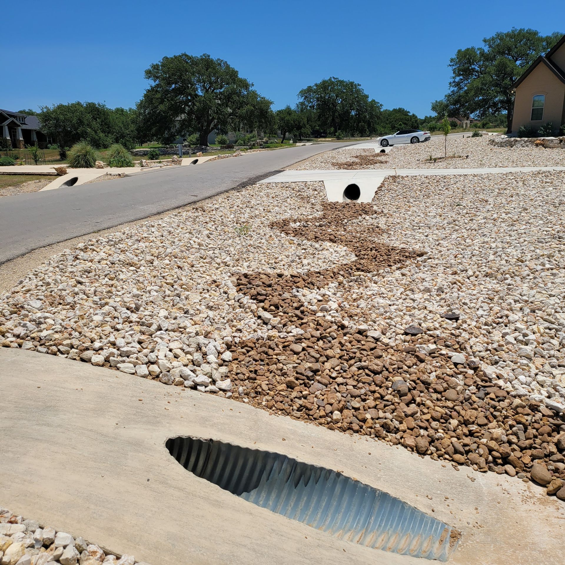 Roadside drainage culvert in a gravel-lined ditch beside a paved street under a clear blue sky