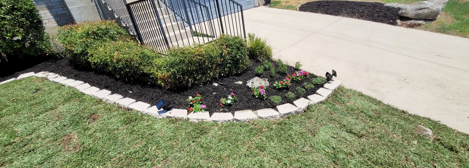 Curved flower bed with white edging, pink blooms, and green shrubs beside a sidewalk and lawn.