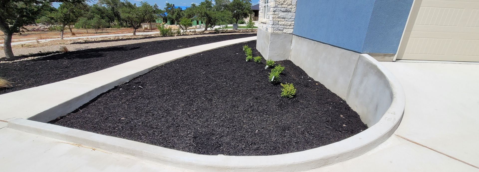 Curved concrete walkway beside a building, bordered by black mulch and small green plants under trees.