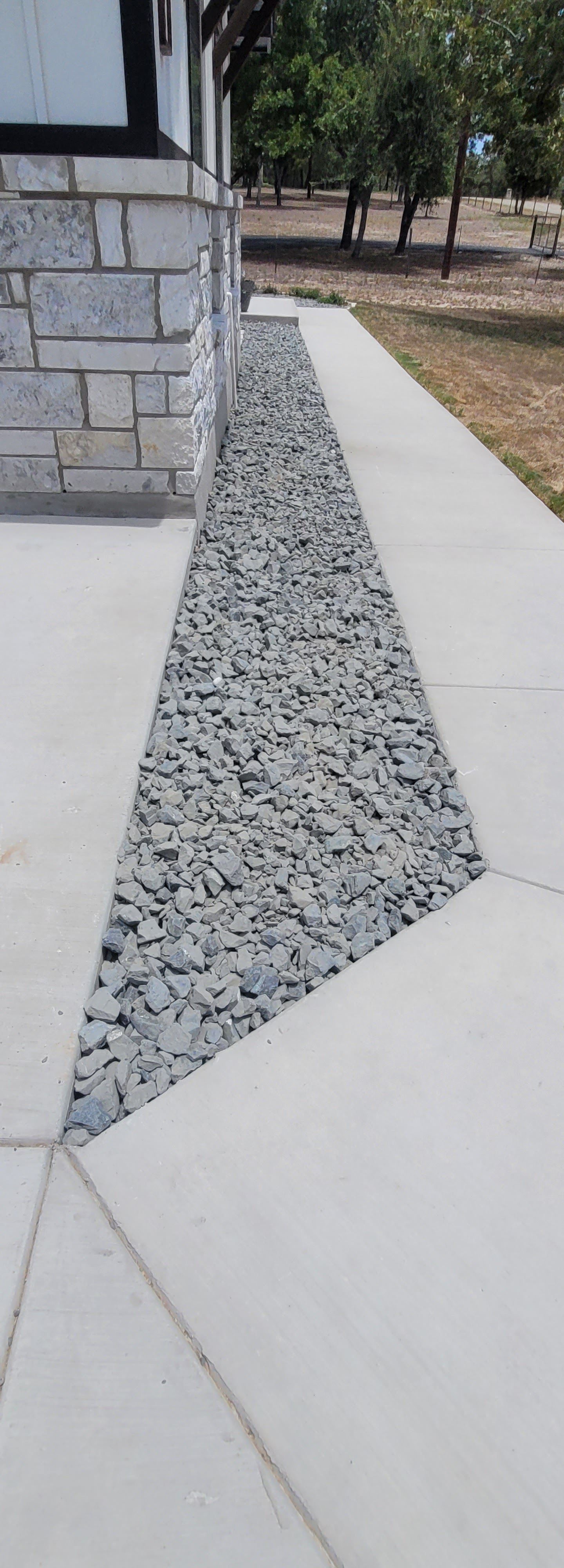 Concrete sidewalk with a gray gravel drainage strip beside a stone wall and trees.