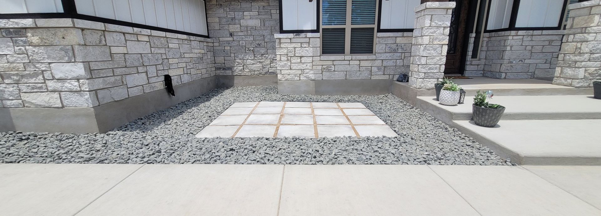 Front entrance of a stone building with a tiled walkway, gravel border, and two small potted plants near the door