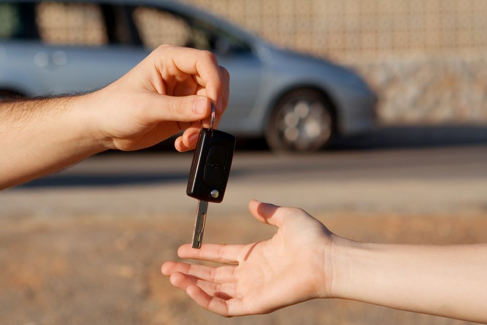 Hands exchanging a car key in front of a blurred parked car outdoors