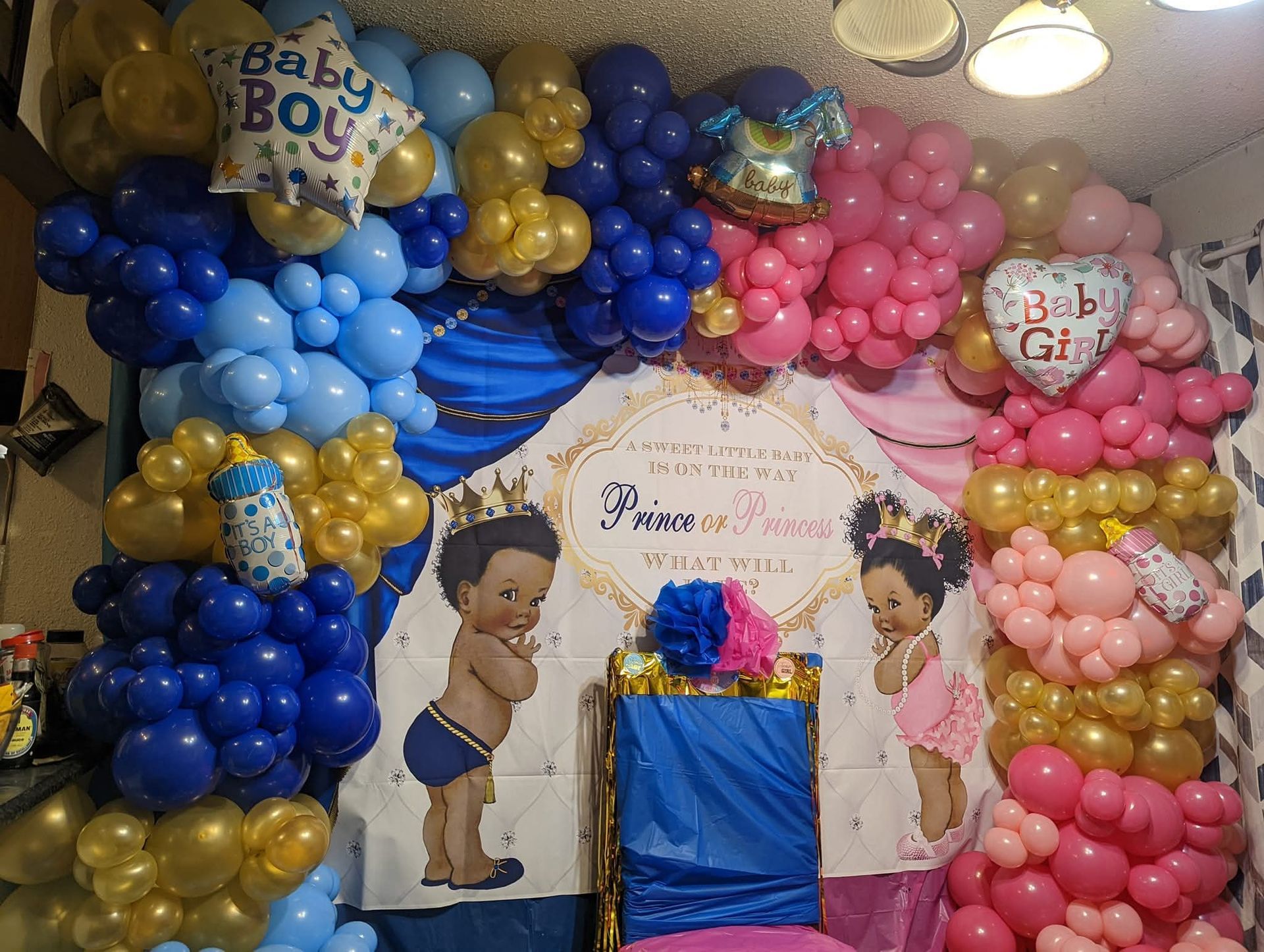 Baby shower backdrop with blue, pink, and gold balloons and cartoon cutouts around a central table.