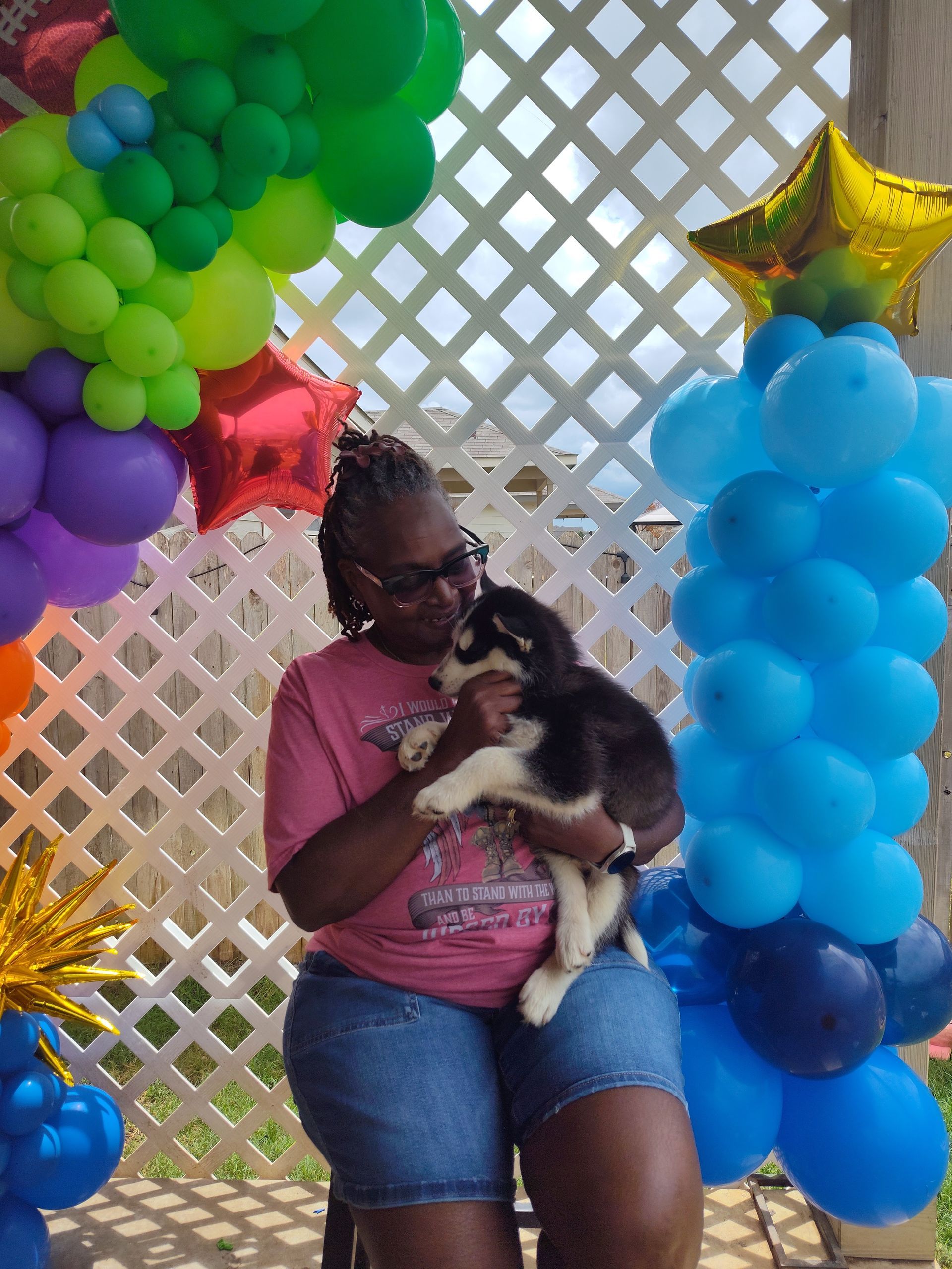 Person holding a black-and-white dog between colorful balloon arches and a lattice backdrop