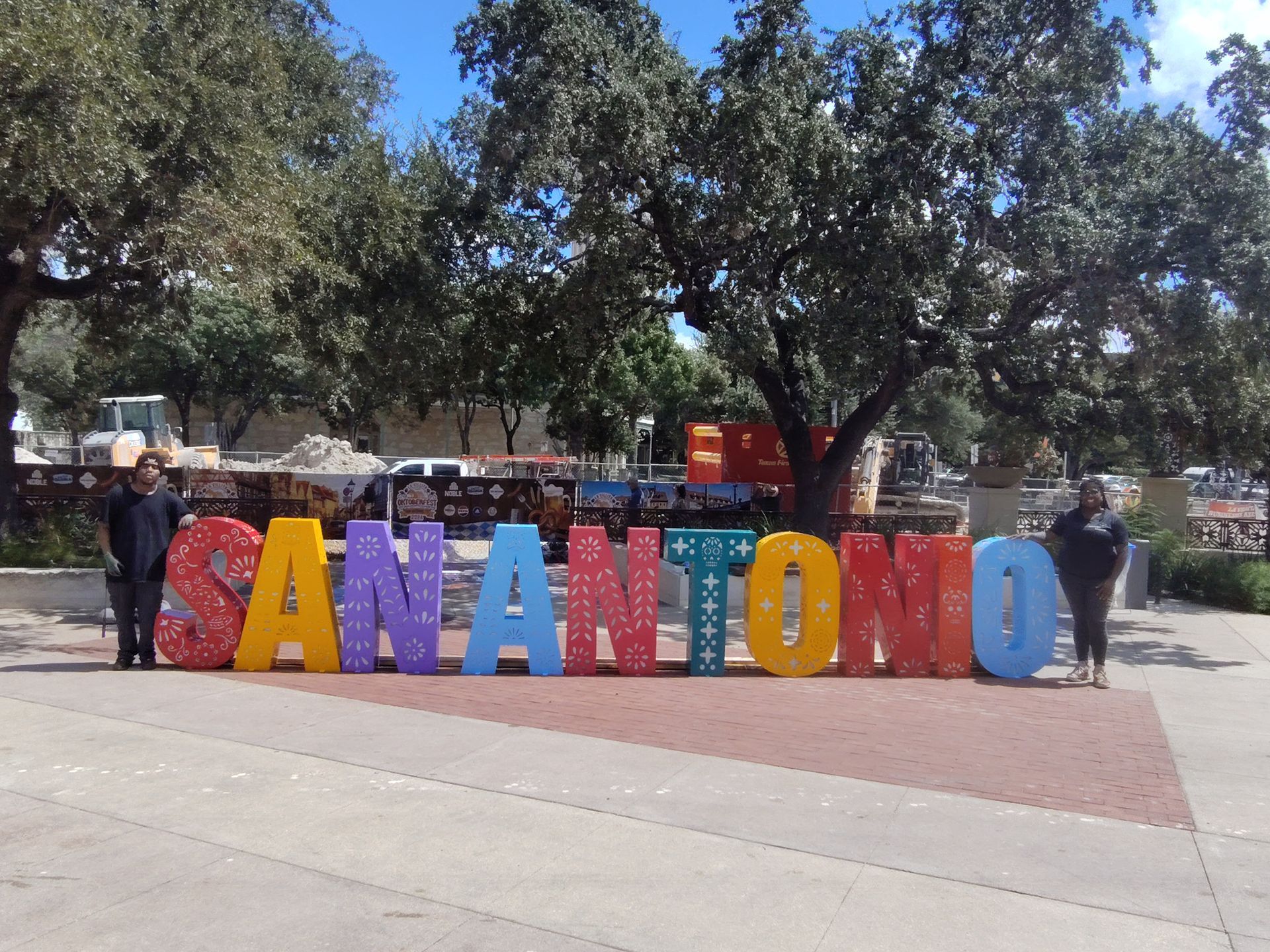 People standing behind colorful large letters spelling “SAN ANTONIO” in a sunny plaza.
