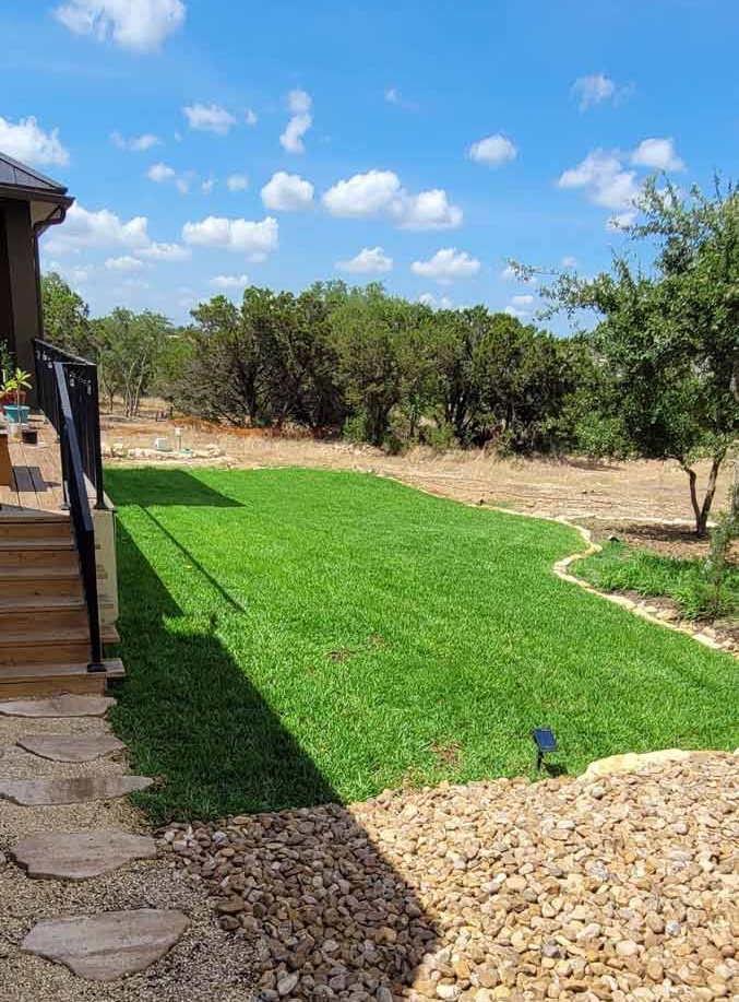 Backyard with green lawn, stone path, gravel border, and trees under a blue sky