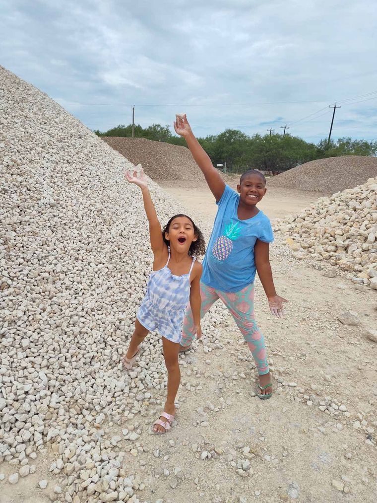 Two smiling children pose with arms raised between large piles of pale rocks outdoors.