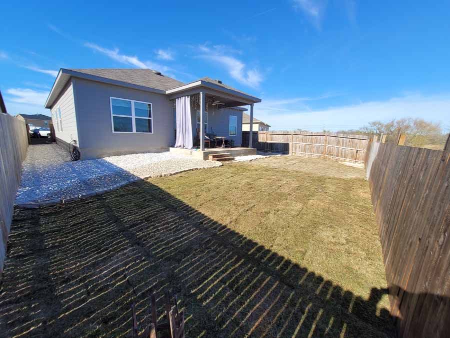 Backyard view of a single-story house with a small porch, grass lawn, and wooden fence under a blue sky