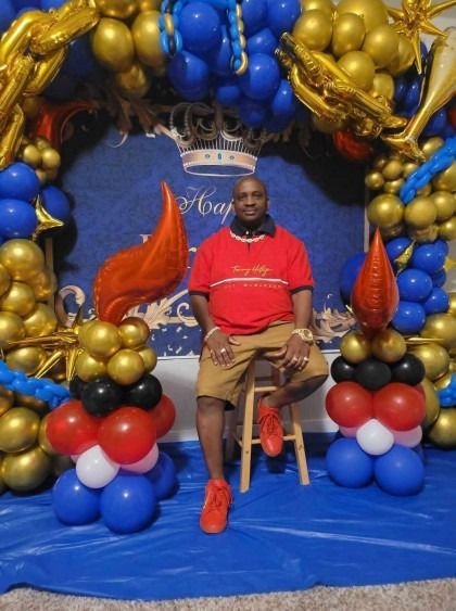 Person seated in front of blue and gold balloon arch, wearing red shirt and shorts at an event