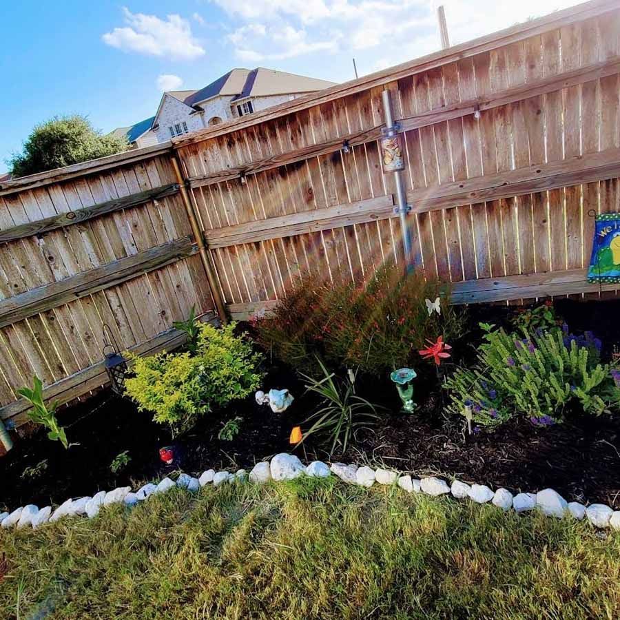 Backyard flower bed with colorful plants bordered by white stones and a wooden fence