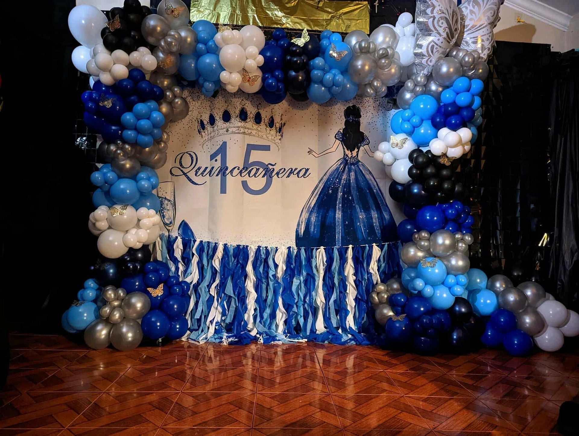 Blue, silver, and black balloon arch framing a decorated stage with a sparkling blue gown backdrop