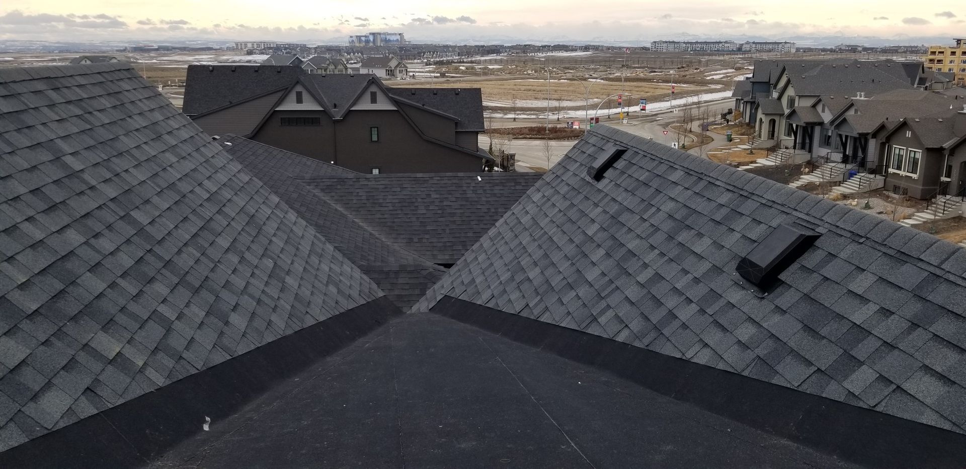 View of a neighborhood from a rooftop. Several dark gray shingled roofs are in the foreground.