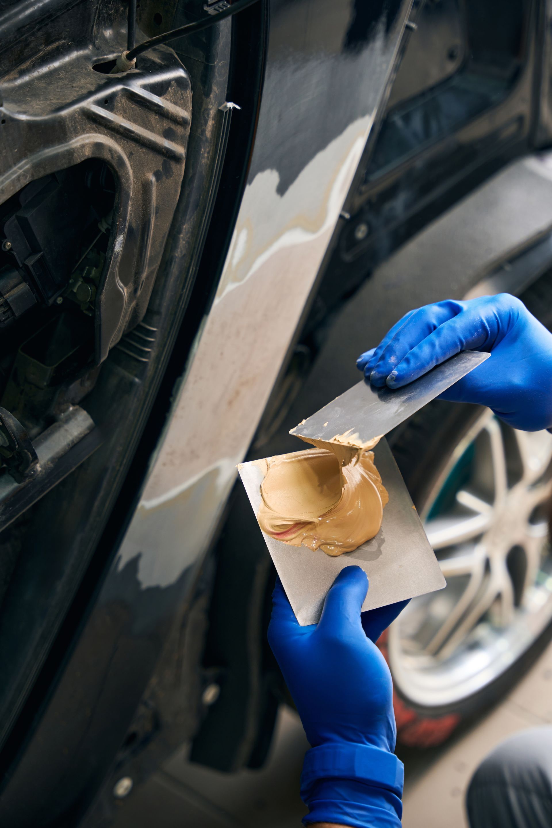 Gloved hands apply tan adhesive to a metal panel beside a car wheel