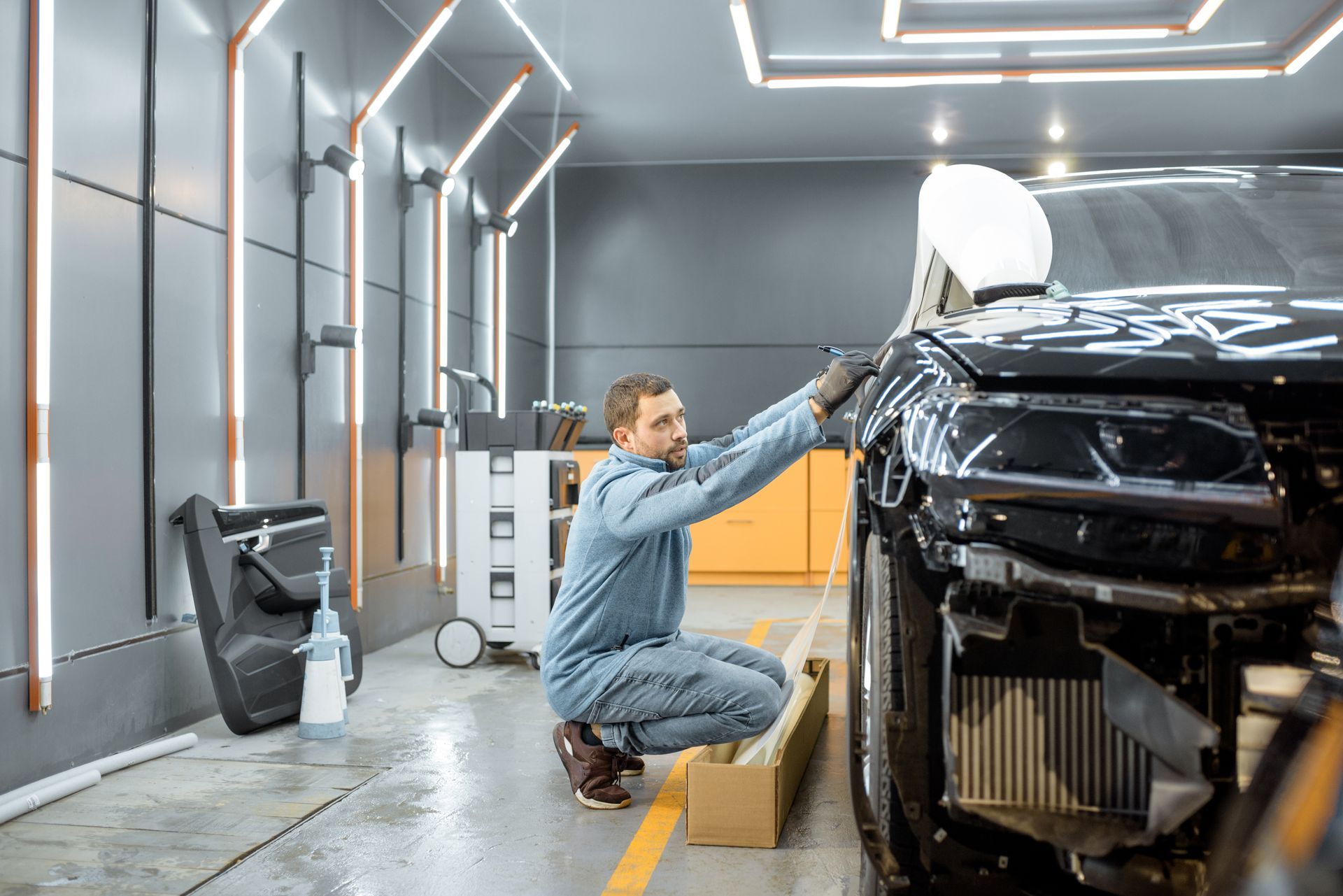 Technician applying protective film onto a black car in an auto body repair shop.