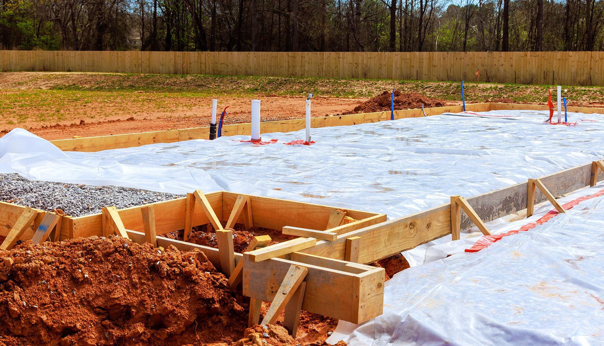 Construction site: wooden foundation formwork, white barrier, exposed pipes, and leveled earth.