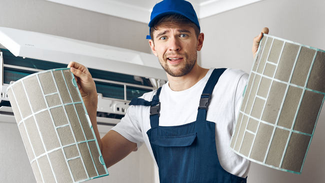 A man is holding two dirty filters in front of an air conditioner.