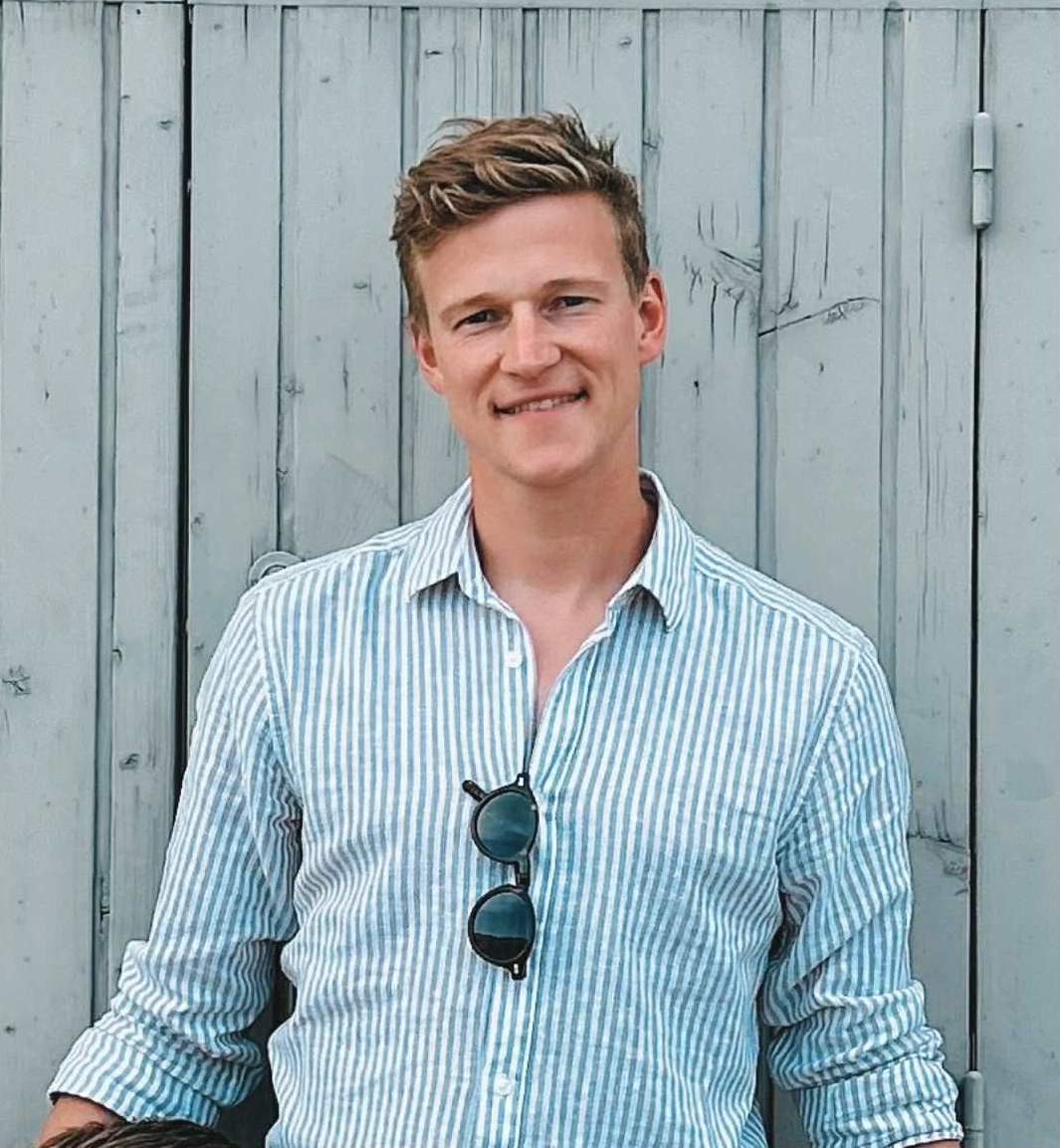 Man with short, light brown hair smiles, wearing a blue striped shirt and sunglasses, in front of a weathered gray wooden wall.