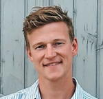 Man with light brown hair smiles, wearing a striped shirt, standing in front of a blue-gray background.