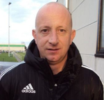 Man with a shaved head wearing a black Adidas jacket, looking at the camera outside.