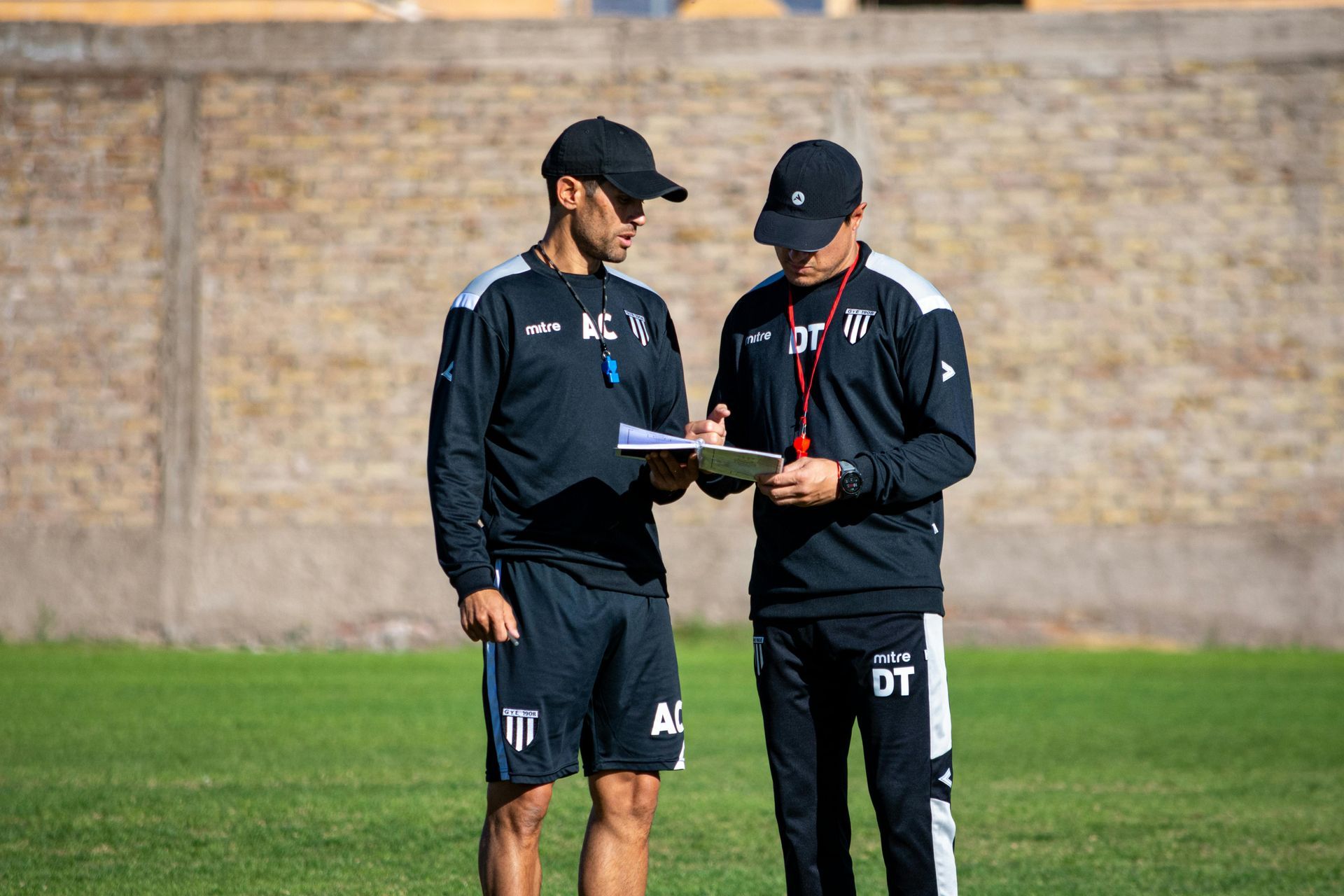 Two soccer coaches in black tracksuits review notes on a green field.