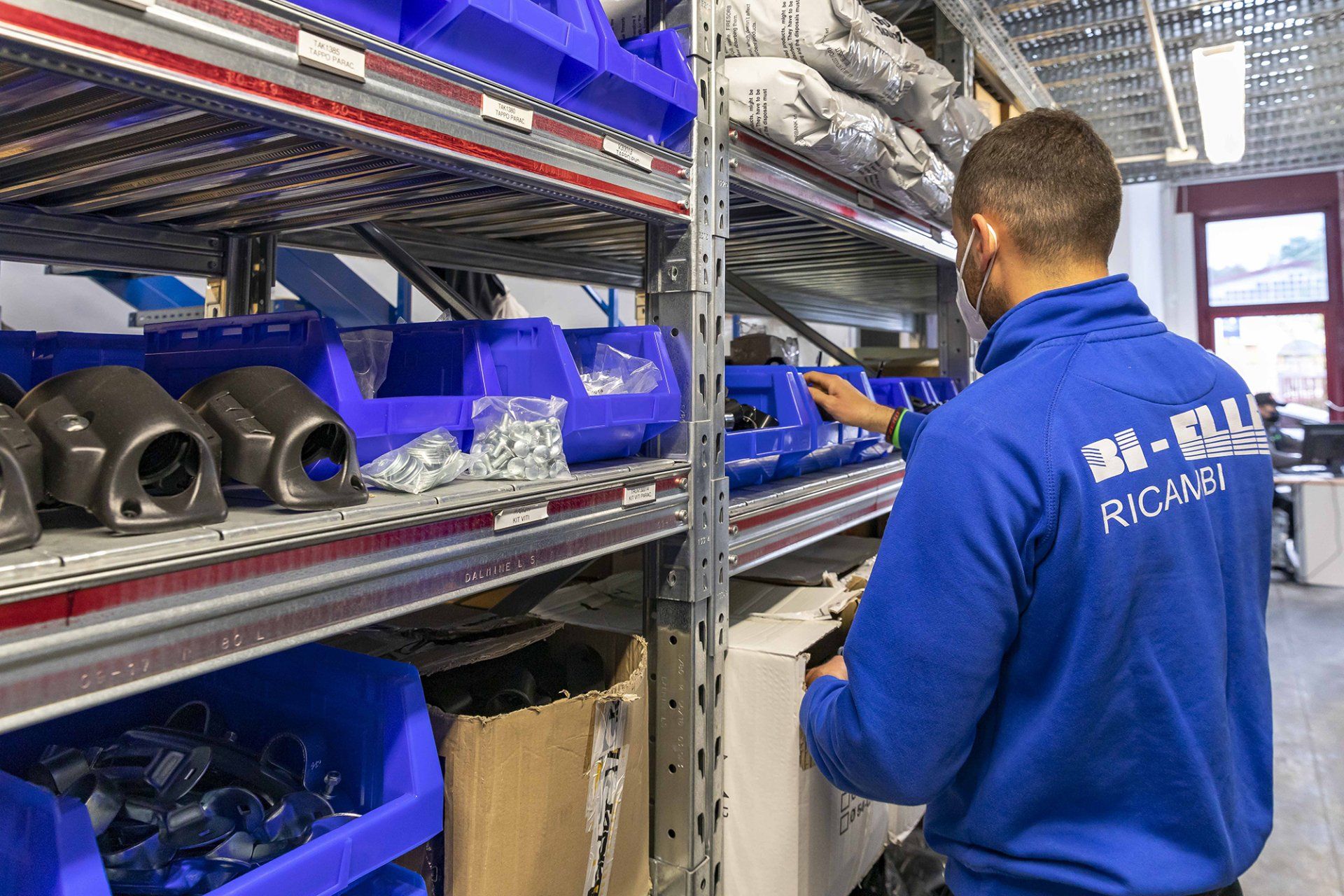 Un operaio con la giacca blu ispeziona dei pezzi su scaffali metallici in un corridoio di un magazzino.