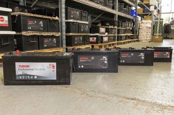 Batterie allineate sul pavimento di un magazzino, davanti a scaffali e scatole impilate.