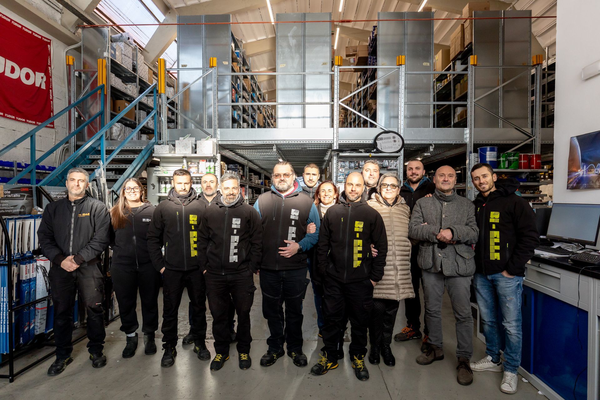 Gruppo di persone in uniforme nera in piedi in un'officina industriale con impalcature metalliche e attrezzature.
