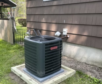 Outdoor air conditioning unit on a concrete pad beside a house with brown siding and a fenced yard.