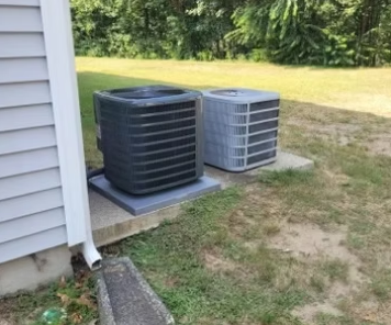 Two outdoor air conditioner units, one black and one gray, beside a house on concrete pads.