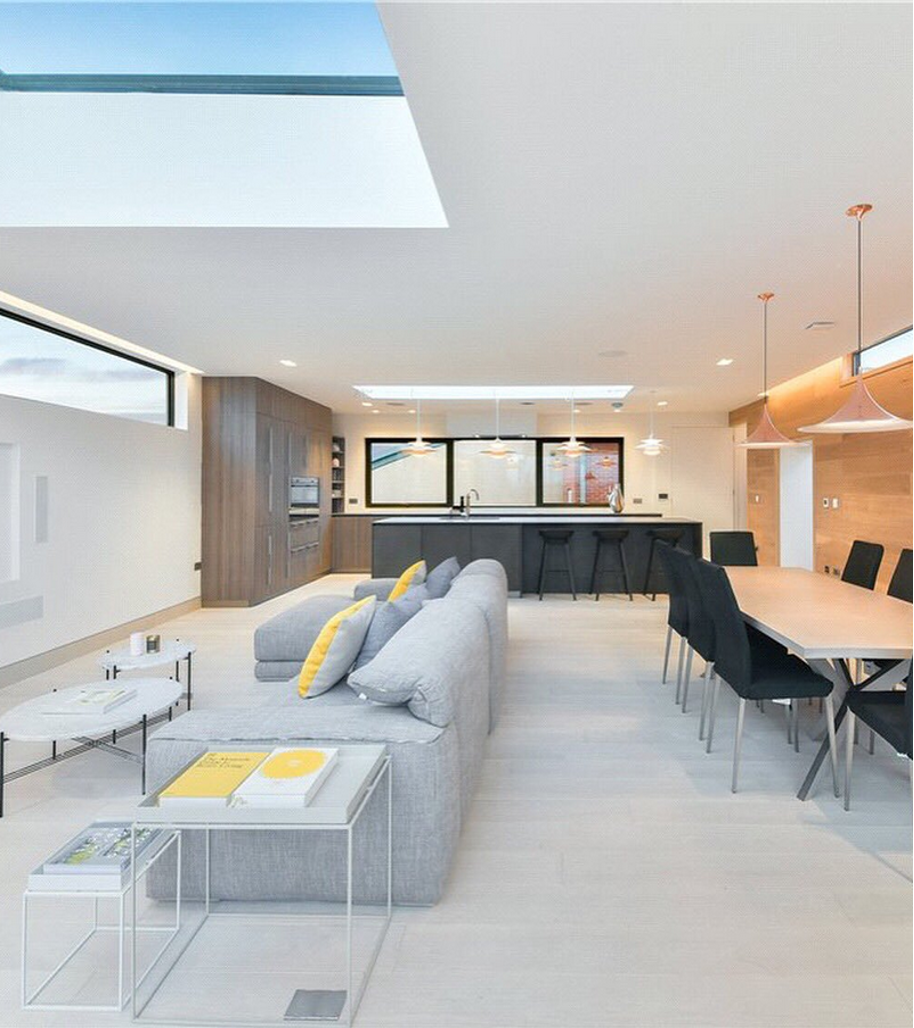 Spacious modern interior with living, dining, and kitchen areas. Features gray sofa, dining table, and skylights.