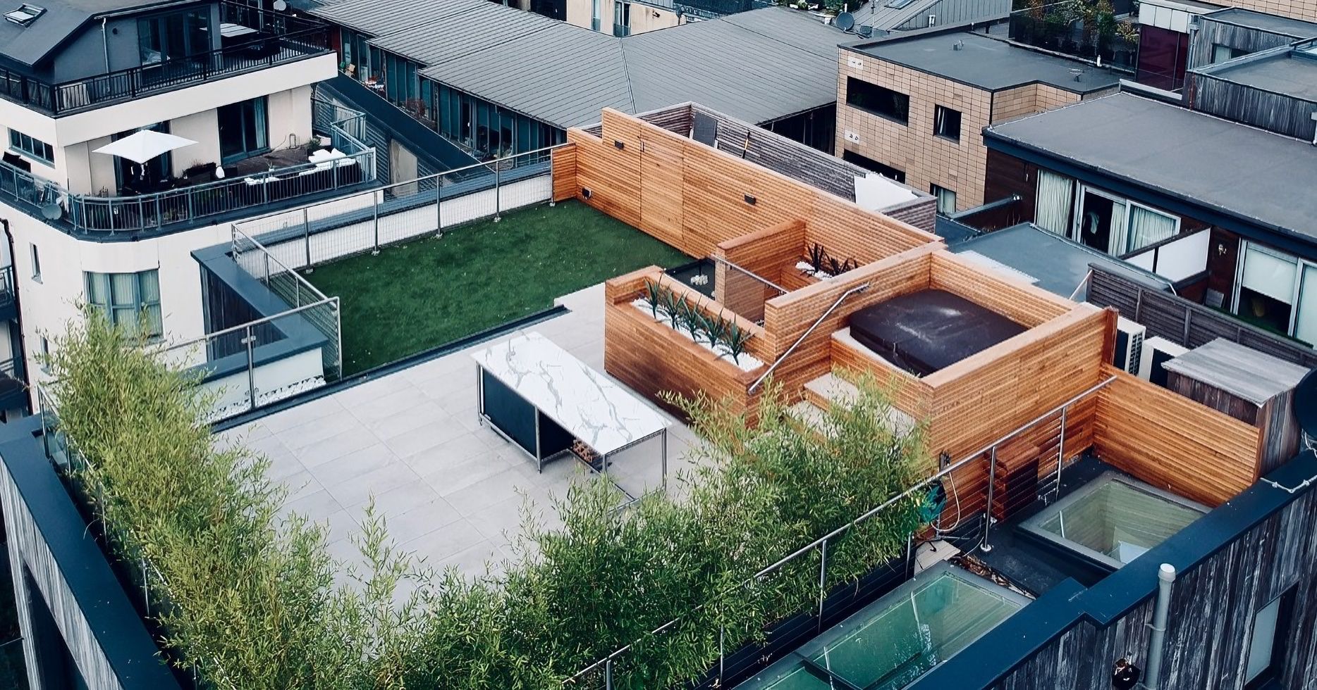 Rooftop patio with wooden structures, green lawn, hot tub, and city views.
