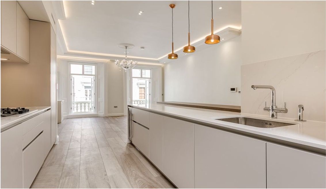 Modern white kitchen with island, wooden floors, pendant lights, and doors to a balcony.