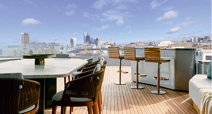 Rooftop deck with seating, bar stools, and a hot tub, overlooking a city skyline under a blue sky.