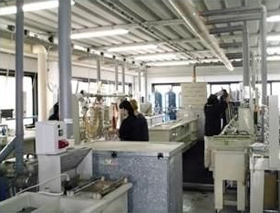 Persone che lavorano in un laboratorio con attrezzature, vari contenitori e ventilazione, vicino alle finestre.