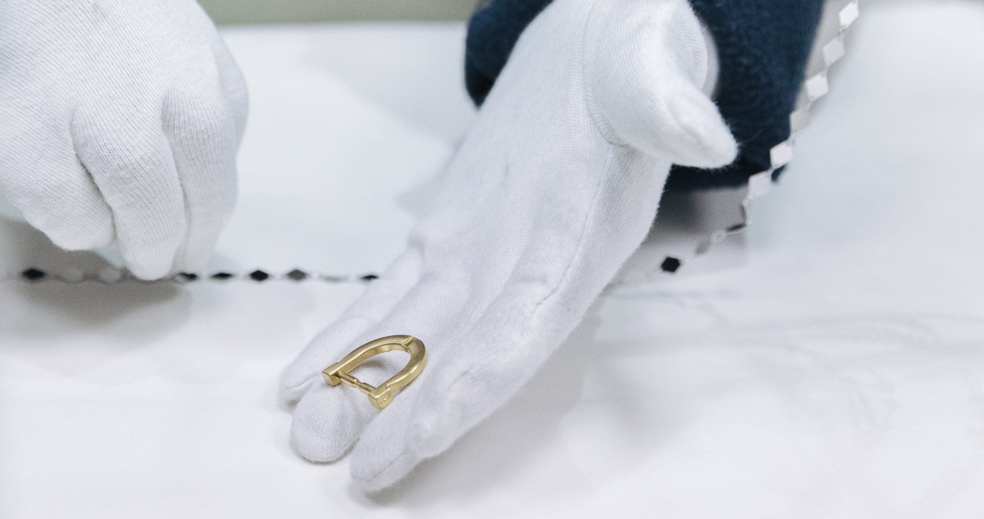 Mani guantate che tengono un anello d'oro su un tessuto bianco, vicino a una linea nera.