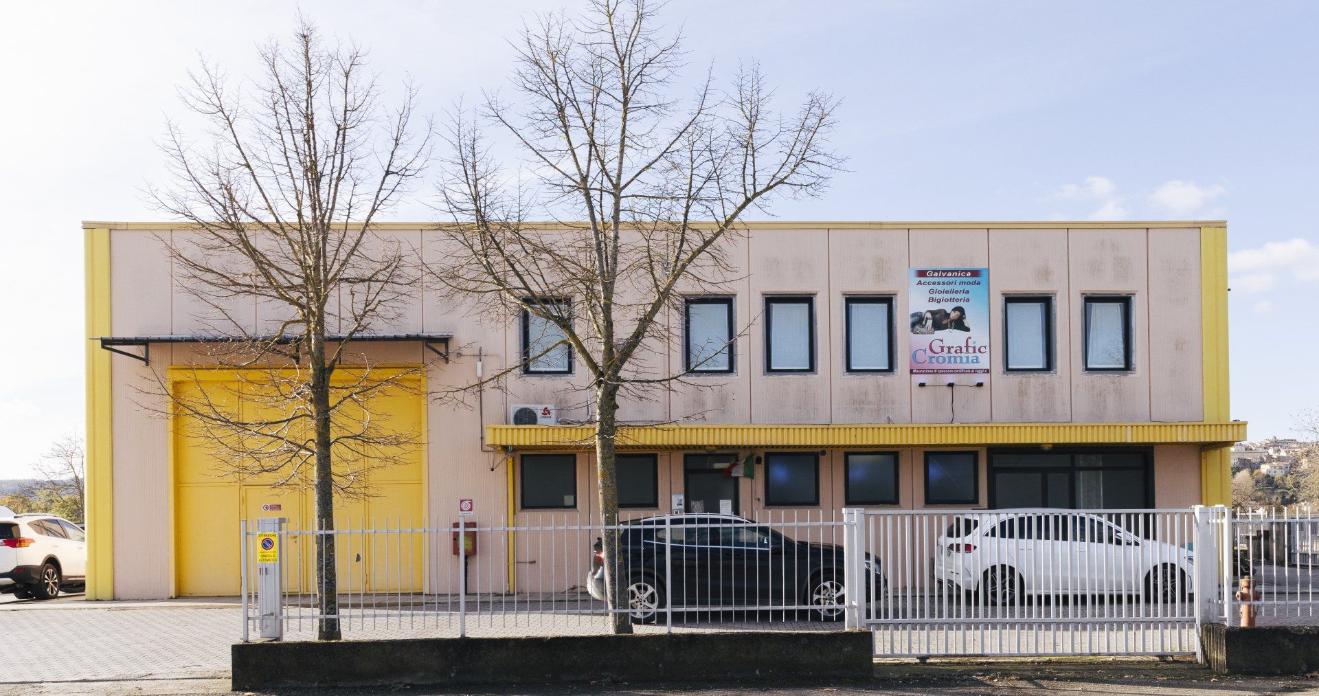 Edificio industriale giallo e marrone chiaro con alberi spogli, automobili e una recinzione metallica.