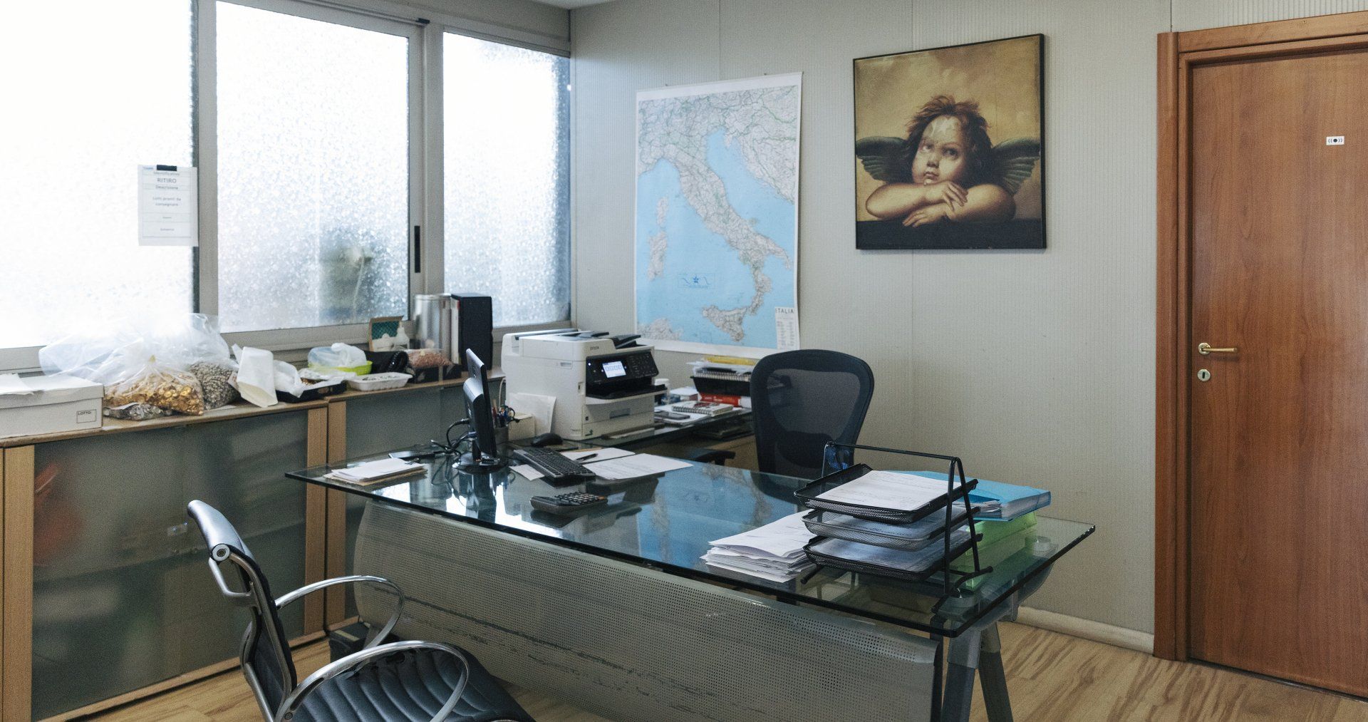 Ufficio con scrivania in vetro, computer, stampante, mappa dell'Italia, quadro e finestra.