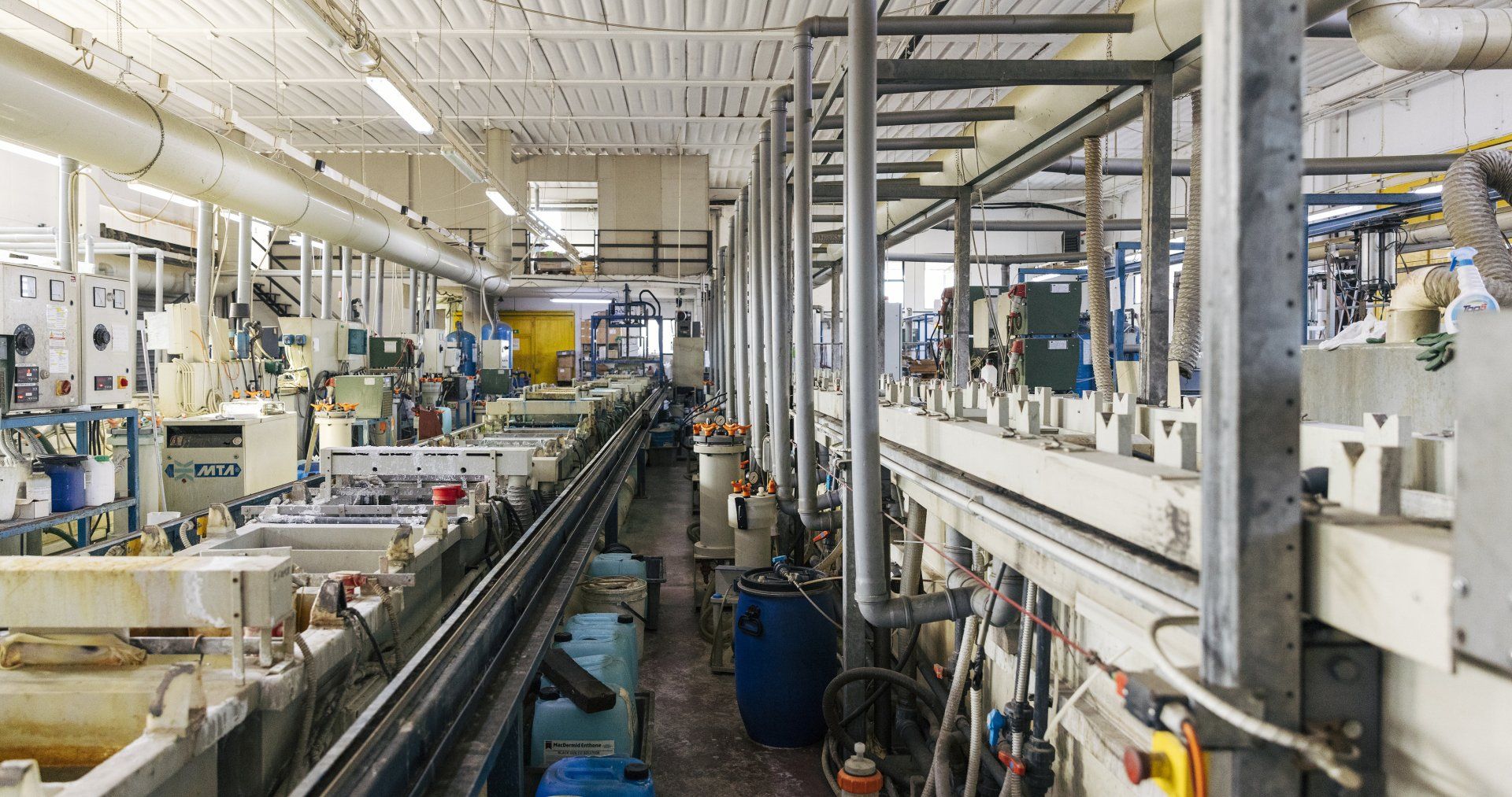 Interno di una fabbrica industriale, con lunghe file di attrezzature, tubi e serbatoi; sono presenti vari colori.